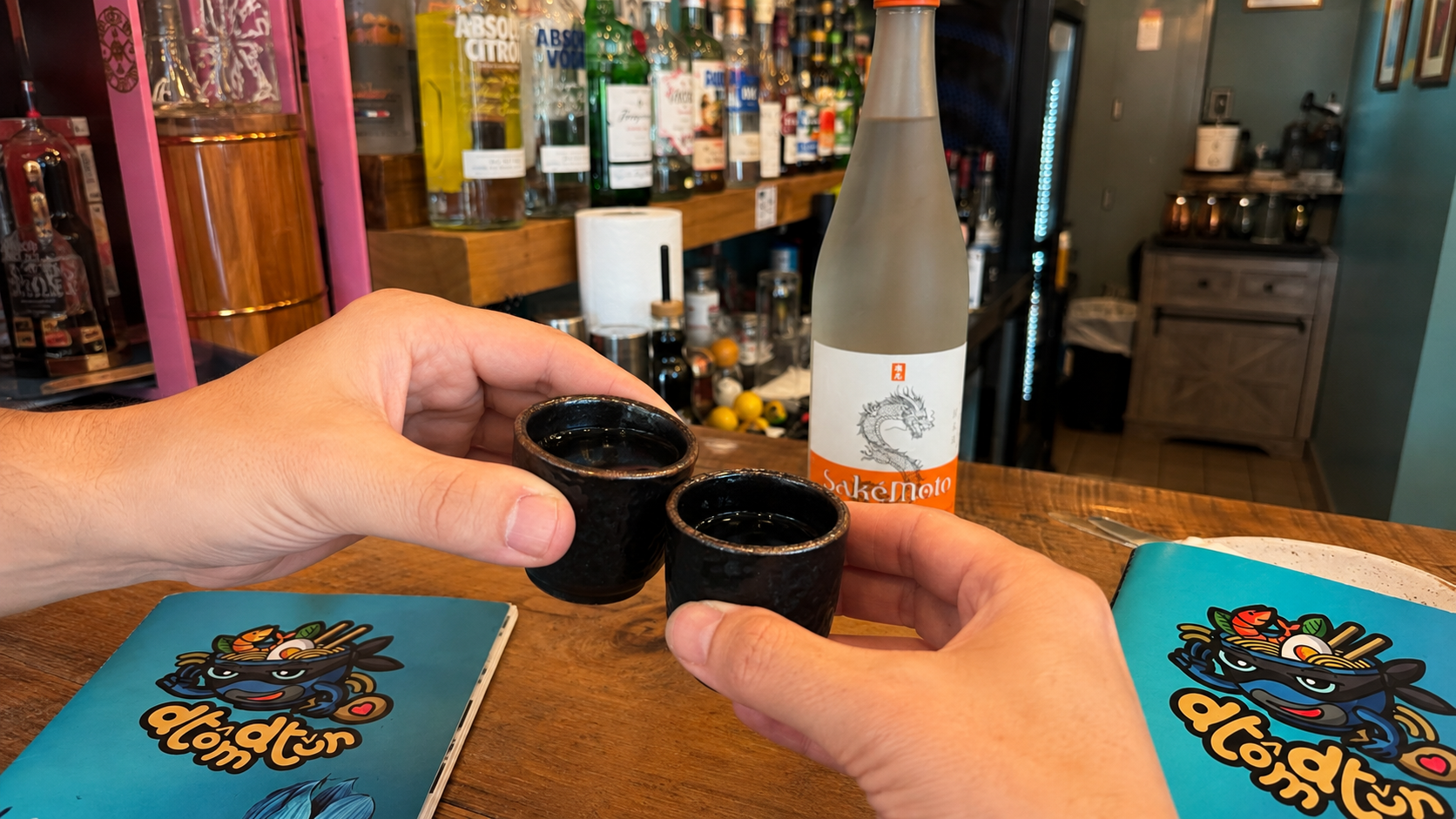 Two people raise black sake cups beside a SakeMoto bottle and Dtom Dtun menus at the Culpeper restaurant.