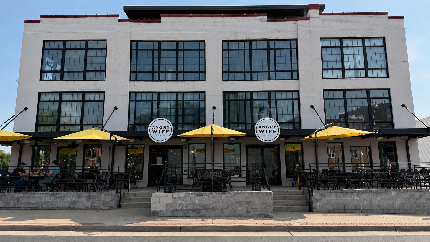 Exterior of Angry Wife Brewhouse at 201 Waters Place in Culpeper, with yellow patio umbrellas and the building's industrial facade.